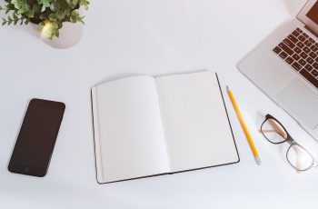 Otwarty notes z ołówkiem, okularami, telefonem i laptopem na białym biurku